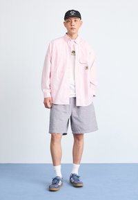 Lyserosa skjorte med knapper, hvit grafisk t-skjorte, gråstripete shorts, blå joggesko, svart caps, satt mot en enkel bakgrunn.