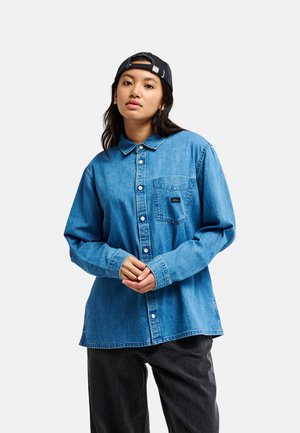 Jeune femme portant une chemise en jean bleu boutonnée, un jean noir et une casquette noire à l'envers, debout devant un fond uni clair.