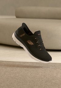 Μαύρο αθλητικό παπούτσι τύπου slip-on με υφή στο επάνω μέρος, ελαφριά λευκή σόλα και μεταλλικές λεπτομέρειες. Διαθέτει διακριτικό γεωμετρικό μοτίβο σε όλο το παπούτσι.