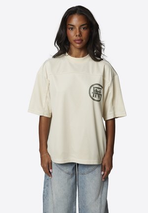 Vrouw met lang donker haar draagt een losse crème kleurige mesh T-shirt met een ronde zwarte logo op de borst en een verwassen, wijd uitlopende jeans, staand en recht vooruit kijkend.
