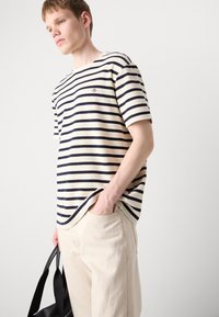 Striped short-sleeve t-shirt in cream and navy, featuring a crew neckline and ribbed texture, paired with light-colored trousers and a black bag.