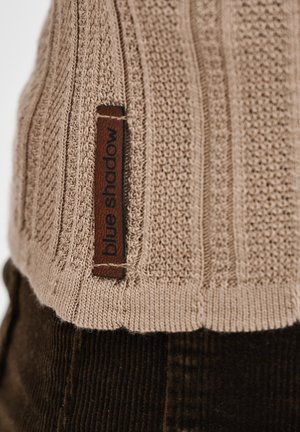 Beżowy sweter dzianinowy z teksturowanym wzorem, z brązową skórzaną metką oznaczoną "blue shadow" na dolnej krawędzi.