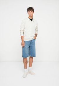 Polo shirt bej deschis cu mânecă lungă, șorturi din denim albastre și pantofi slip-on crem. Șorturile sunt de lungime până la genunchi, cu o croială lejeră și un stil casual.