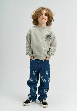 Szürke kapucnis pulóver grafikai dizájnnal, kék farmerrel, amely világoskék mintákkal rendelkezik. Fekete sportcipők egészítik ki az öltözetet.