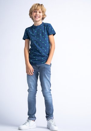 Marineblauw t-shirt met korte mouwen en kleine groene patronen, gecombineerd met lichtblauwe jeans en witte sneakers. Model staat in een ontspannen houding.