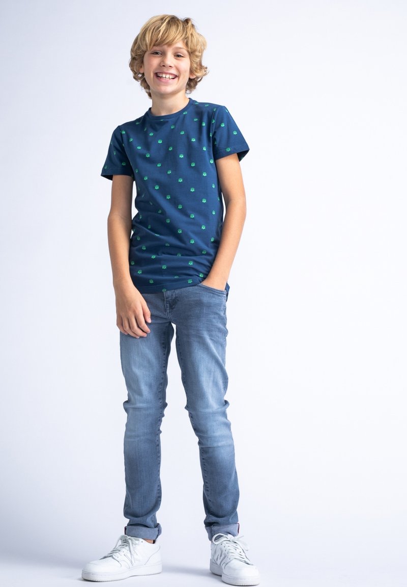 Tricou de navy cu mânecă scurtă, cu modele mici verzi, asortat cu blugi deschiși și teniși albi. Modelul stă într-o postură relaxată.