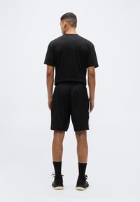 Άνδρας που στέκεται με την πλάτη προς τα εμπρός, φορώντας μαύρο μπλουζάκι, μαύρο αθλητικό σορτς με άσπρες πλευρικές ρίγες, μαύρες κάλτσες και μαύρα αθλητικά παπούτσια.