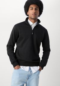 Black zip-up sweatshirt with a high collar, featuring a contrasting horizontal seam; worn over a white collared shirt and blue denim.