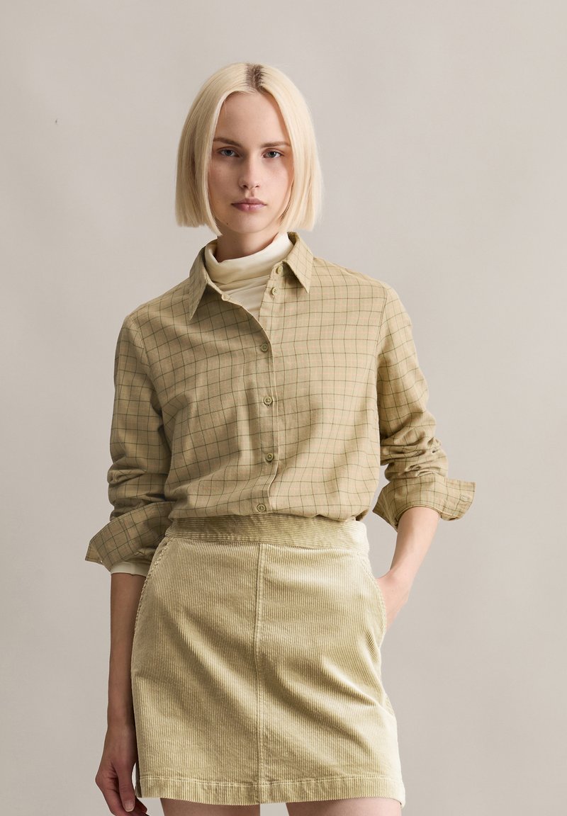 Camicia a quadri marrone chiaro con bottoni, maniche arrotolate, abbinata a un dolcevita color crema e a una gonna in velluto a coste chiaro.