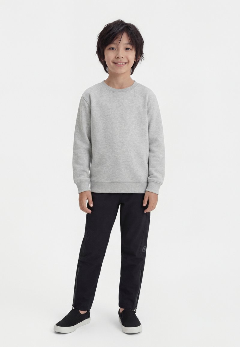 Niño sonriente de pie, con sudadera gris claro, pantalones negros con cremallera en los tobillos y zapatos negros sin cordones con suelas blancas.