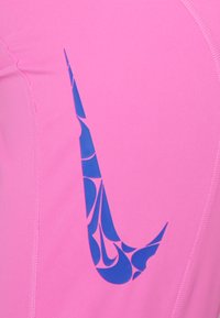 Růžový sportovní top s modrým logem Nike Swoosh, který obsahuje geometrický vzor, vyrobený z elastického materiálu, navržený pro pohodlí a mobilitu.