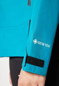 Tirkizna GORE-TEX jakna s čičak manžetama. Glatka tekstura s elegantnim dizajnom i reflektirajućim detaljima. Prikaz detalja na rukavu.