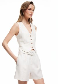Vrouw in een mouwloze beige knopenvest en bijpassende shorts met riem, staand met één hand in de zak tegen een witte achtergrond.