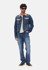 Chaqueta de mezclilla con cuello y bolsillos beige, superpuesta sobre una camisa clara, combinada con jeans azules descoloridos y zapatillas oscuras. Gafas de sol rectangulares.
