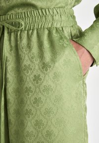 Svetlo zelena tkanina z embosiranim cvetličnim vzorcem, elastičen pas z vrvico in stransko žepom, ki je viden na oblačilu.