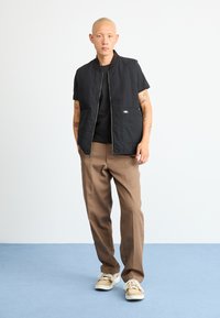 Črna prešita telovnik z zadrgo spredaj in dvema sprednjima žepoma, nošen preko črnega majice, v kombinaciji s svetlo rjavimi hlačami in večbarvnimi športniki.