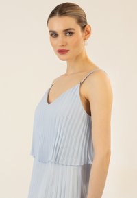 Helles blaues, plissiertes Kleid mit V-Ausschnitt, geschichteten Einschnitten und silbernen Akzenten an den Trägern. Glatte Stoffoberfläche, verstellbare Schulterträger.