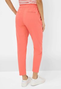 Koraalkleurige slim-fit broek met een elastische tailleband, voorzien van twee achterzakken en taps toelopende benen, gedragen met witte sneakers. Gladde textuur.