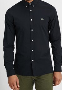 Svart skjorta med långa ärmar, vita knappar och en liten grön logotyp på bröstet. Tyget verkar slätt och lätt.