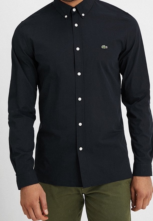 Svart skjorta med långa ärmar, vita knappar och en liten grön logotyp på bröstet. Tyget verkar slätt och lätt.