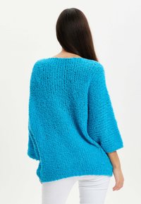 Loose-fitting, bright blue knitted sweater with a textured surface, three-quarter sleeves, and a rounded neckline. White jeans visible below.