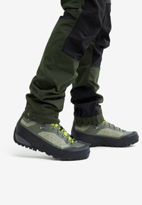 Legs wearing green and black pants tucked into green and black hiking boots with yellow laces on white background.