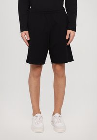 Pantalones cortos de algodón negros con cintura elástica, bolsillos laterales y corte recto. Llevado con zapatillas blancas. Diseño simple y versátil.