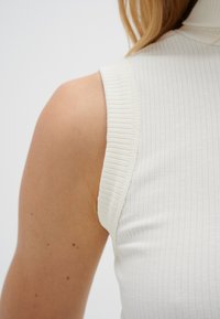 Vit ribbad ärmlös topp med hög krage, med texturerad tyg och en figurnära design som visar mjuka axlar och hudton.
