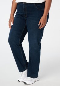 Donkerblauwe denim jeans met een rechte pijp, voorzien van standaardzakken, zichtbare stiksels en gedragen met witte sportschoenen.