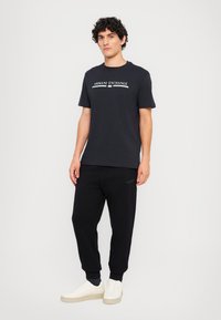 Svart bomull T-skjorte med hvit "Armani Exchange" logo; kombinert med svarte joggebukser og kremfargede sneakers. Enkel design, avslappet passform.