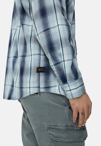 Blau kariertes Hemd mit weißen und dunkelblauen Quadraten, mit Knopfmanschetten und einem Etikett mit der Aufschrift "Made for Adventures". Getragen zu grauen Cargohosen.