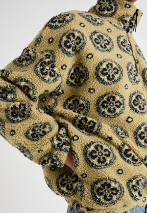 Beżowa kurtka sherpa z polaru z czarnymi i ciemnozielonymi okrągłymi wzorami kwiatowymi, wysokim kołnierzem i rękami w bocznych kieszeniach.