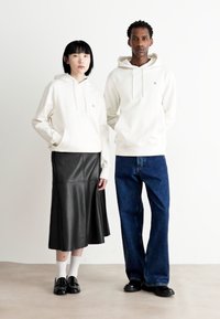 Due modelle indossano una felpa bianca con cappuccio, abbinata a una gonna di pelle nera e jeans blu. I dettagli includono una tasca a marsupio e un taglio morbido.