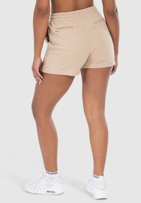 Pantalones cortos de algodón beige con cinturilla elástica, que cuentan con dos bolsillos traseros y una discreta etiqueta de logo, combinados con zapatillas deportivas blancas.
