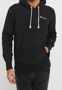 Sudadera negra de tejido suave con un bolsillo canguro frontal, capucha ajustable con cordón y logo bordado en el pecho.