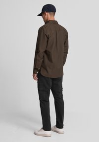 Brown button-up shirt with a back pleat, paired with black cargo pants and white sneakers, worn with a navy cap.