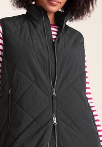 Woman wearing a black quilted sleeveless vest over a pink and white striped long-sleeve shirt, with two zippered pockets visible.