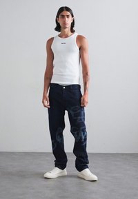Tricou alb fără mâneci, blugi închiși la culoare cu un imprimeu grafic și adidași albi. Ținuta are un design ajustat cu culori contrastante.