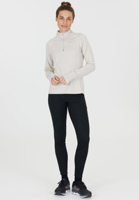 Lys beige langærmet top med halv-lynlås og tommelfingerhuller, parret med sorte tætsiddende bukser. Har en glat tekstur og atletisk design.