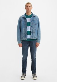 Denim jacket in light blue with front pockets, layered over a striped green and light blue sweatshirt, paired with dark jeans and black sneakers.