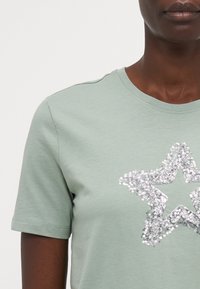 Mintgrønn t-skjorte med korte ermer og et stort sølvfarget paljettstjernesdesign i sentrum. Glatt tekstur og klassisk passform.