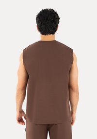 Homme aux cheveux noirs courts et bouclés, portant une chemise marron sans manches et un pantalon marron assorti, vu de dos.