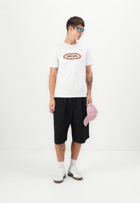 Hvid bomulds t-shirt med et flerfarvet "RIPCURL" logo, sorte shorts, hvide sneakers og holder en pink baseball cap. Ensfarvet baggrund.