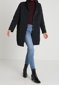 Sort quiltet frakke med høj krave og knapper foran. Båret over en vinrød rullekrave, kombineret med lyseblå jeans og sorte ankelstøvler.