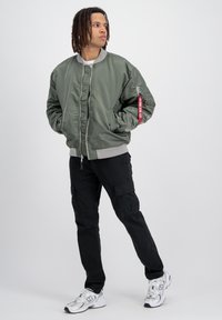 Jachetă bomber de culoare verde măsliniu cu buzunar cu fermoar și etichetă roșie, asortată cu pantaloni cargo negri și adidași albi. Textură netedă, croi lejer.