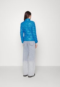 Kobieta stojąca tyłem, ubrana w jasnoniebieską pikowaną kurtkę i szerokie, jasnoniebieskie, wyblakłe dżinsy na jednolitym tle.