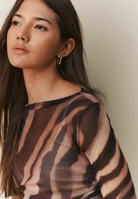 Tunique à manches longues en tissu transparent avec un motif abstrait marron et crème, un col rond et une texture lisse. Boucles d'oreilles en or en anneau en guise d'accents.