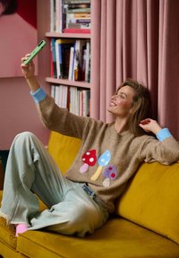 Beżowy sweter z kolorowymi aplikacjami grzybów, jasnoniebieskie mankiety, w połączeniu z luźnymi jasnymi dżinsami. Żółta sofa w przytulnym wnętrzu.