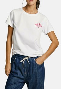 Camiseta blanca de algodón con mangas cortas, con un logo floral rosa en el pecho. Combinada con pantalones de mezclilla azul oscuro con cordón.
