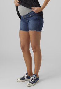 Jeans-Maternity-Shorts mit fransigem Saum, ausgestattet mit einer taillierten Taille und einem leicht lockeren Design. Kombiniert mit marineblauen Sneakers.
